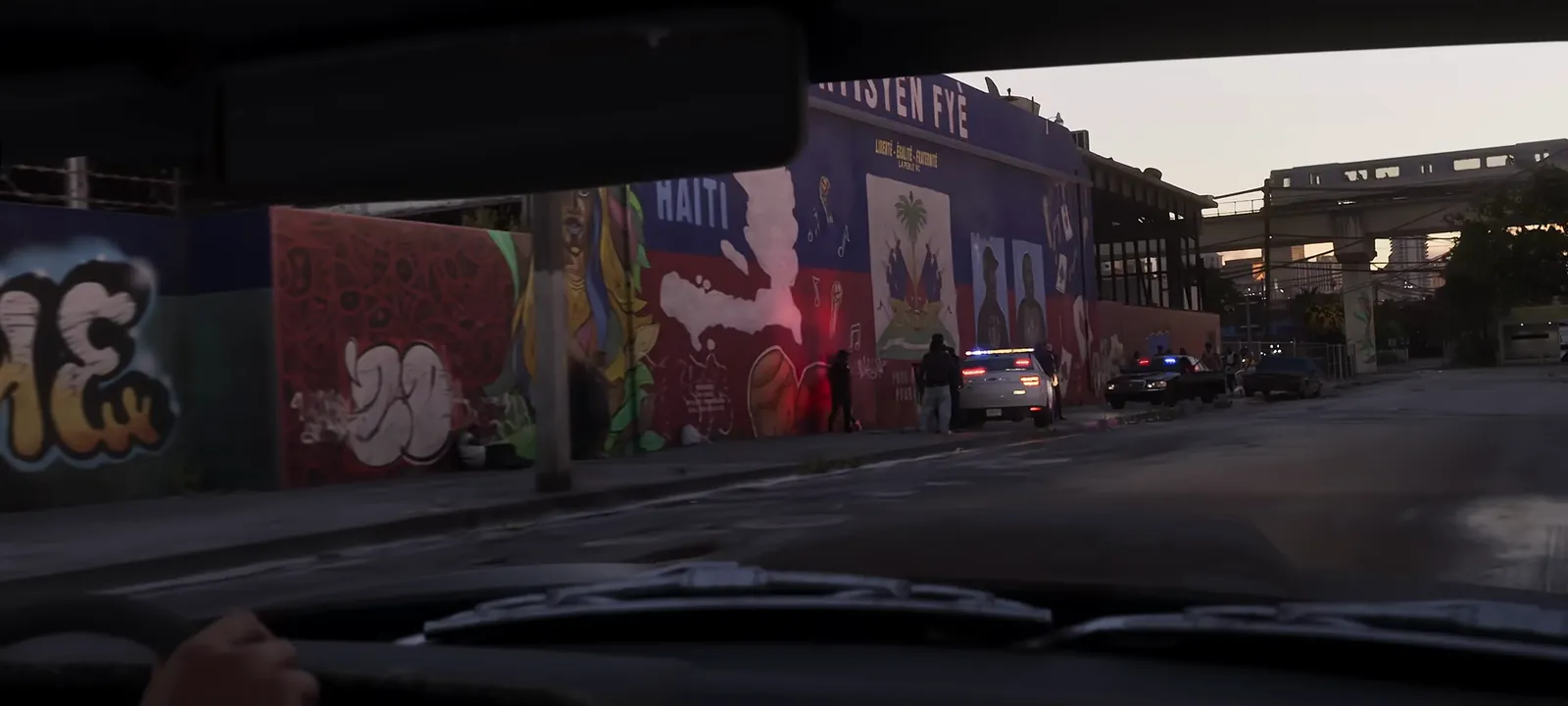 Little Haiti Cultural Complex and Caribbean Marketplace in GTA VI: Driving through La Perle past Haitian-flag murals and a police scene, with an elevated rail line ahead.